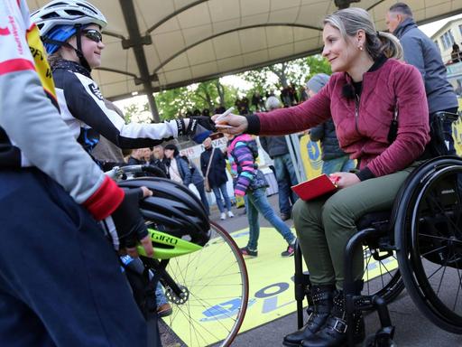Kristina Vogel sitzt im Rollstuhl und übergibt einem Kind in Radkleidung ein Autogramm.