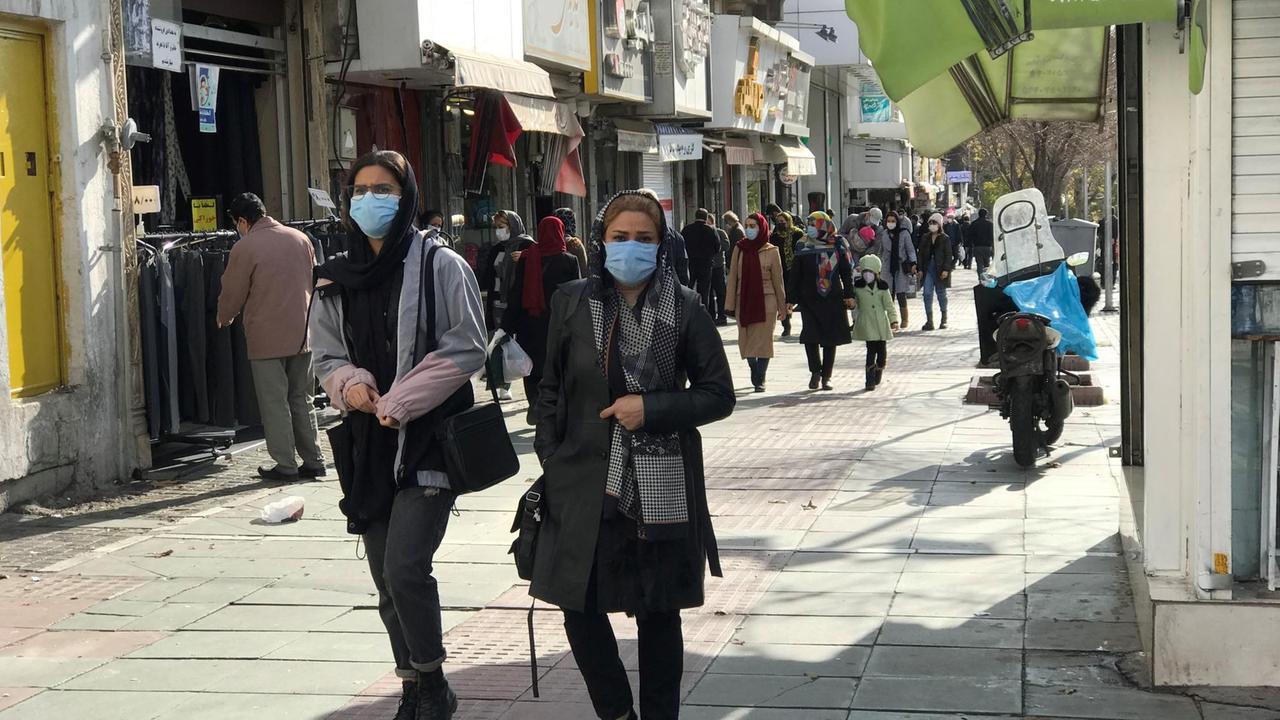 Zwei Frauen mit hellblauem Mundschutz schlendern durch die sonnige Fußgängerzone von Teheran.