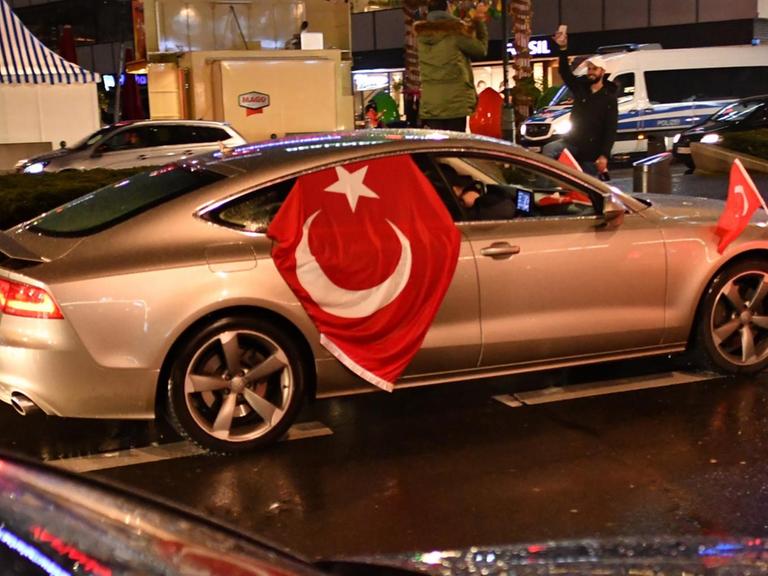 Sie sehen ein silbernes Auto mit einer Türkeifahne in Berlin. Es ist Nacht.