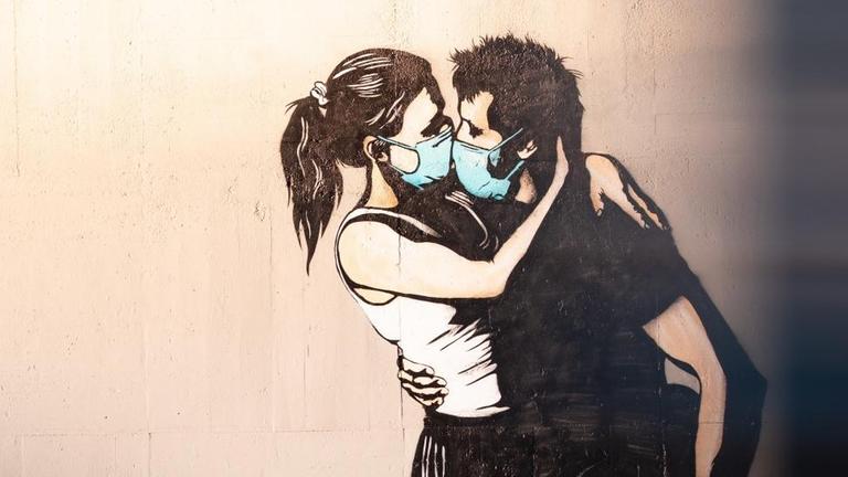 Graffiti an einer Wand: Ein junges Liebespaar mit Coronamasken umarmt und küsst sich. Graffiti an einer Wand: Ein junges Liebespaar mit Coronamasken umarmt und küsst sich.