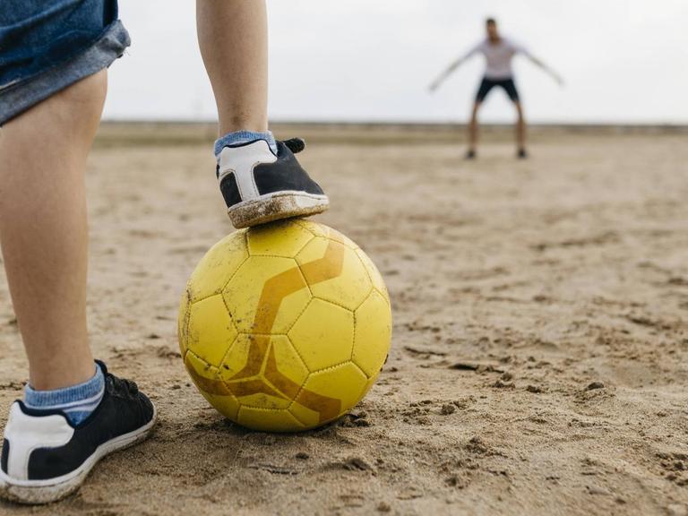 Die Beine eines Jungen mit Fußball, vor einem Tor mit Torwart
