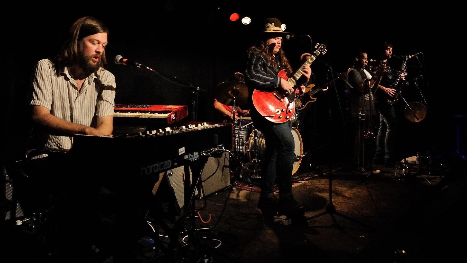 Musiker stehen auf einer Bühne und spielen, in der Mitte ein junger, langhaariger Mann an einer roten Gitarre. Musiker stehen auf einer Bühne und spielen, in der Mitte ein junger, langhaariger Mann an einer roten Gitarre.