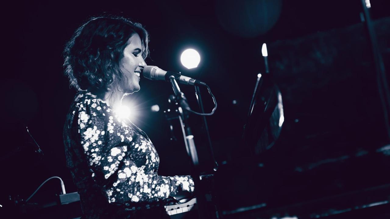 Eine Frau mit dunklen Locken und geblümtem Kleid spielt auf dunkler Bühne Keyboard und singt lächelnd in ein Mikrofon. Hinter ihr leuchten zwei Scheinwerfer.