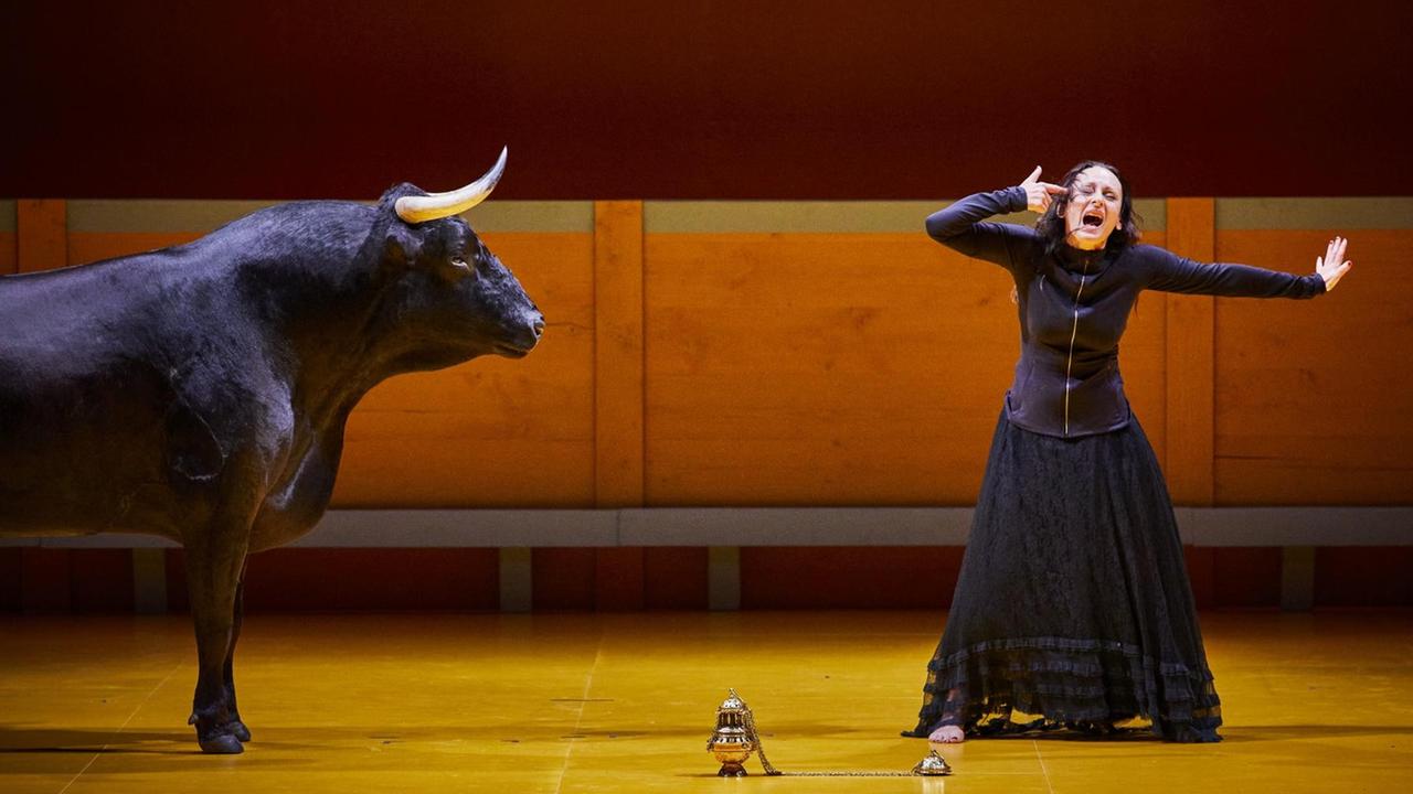 Szenenbild aus der Inzenierung "Liebestod" der katalanischen Regisseurin Angélica Liddell. Eine schreiende Frau im schwarzen Kleid steht neben einem Stier.