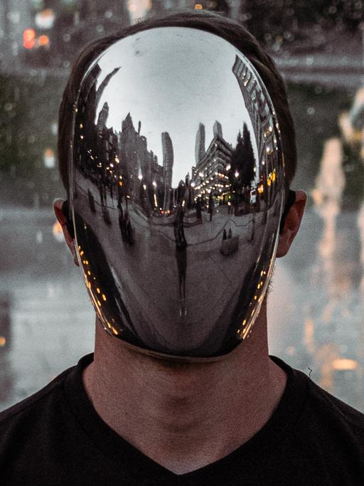 Ein Mann mit verspiegelter Maske steht auf einem Platz mit Hochhäusern.