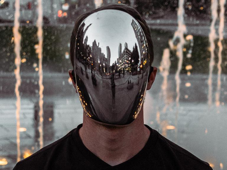 Ein Mann mit verspiegelter Maske steht auf einem Platz mit Hochhäusern.