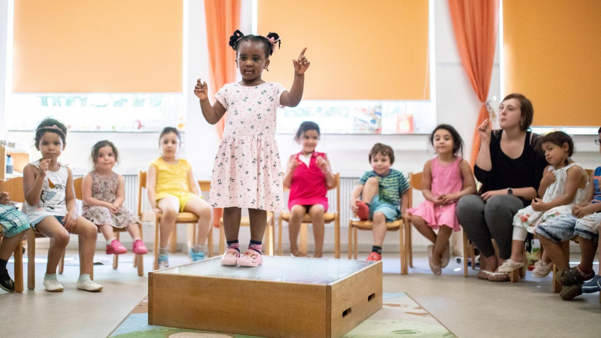 Ein kleines Mädchen steht in einem großen Raum auf eine Podest und macht Gebärden. Um sie herum sitzen viele andere Kinder und gucken ihr dabei zu. Ein kleines Mädchen steht in einem großen Raum auf eine Podest und macht Gebärden. Um sie herum sitzen viele andere Kinder und gucken ihr dabei zu.