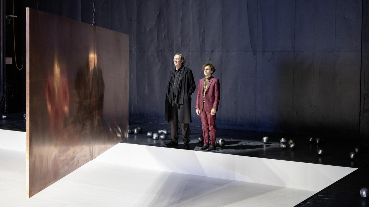 Ein älterer und ein junger Mann stehen am Rande einer Theaterbühne und schauen auf Abbilder ihrer selbst, die sich verzerrt von einer in der Mitte des Raumes hängenden Metallplatte widerspiegeln. Ein älterer und ein junger Mann stehen am Rande einer Theaterbühne und schauen auf Abbilder ihrer selbst, die sich verzerrt von einer in der Mitte des Raumes hängenden Metallplatte widerspiegeln.