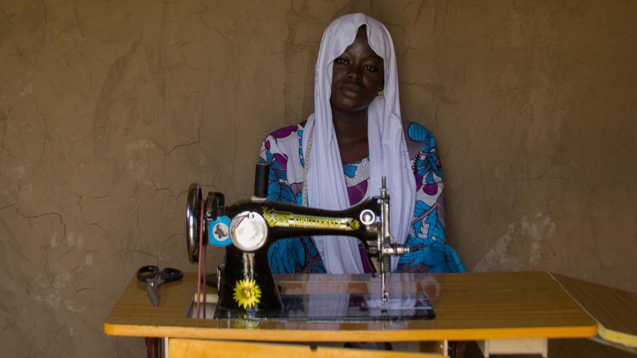 Eine junge afrikanische Frau mit weißem Kopftuch und buntem Kleid sitzt vor einer altmodischen Nähmaschine, die auf einem Klapptisch steht. Eine junge afrikanische Frau mit weißem Kopftuch und buntem Kleid sitzt vor einer altmodischen Nähmaschine, die auf einem Klapptisch steht.