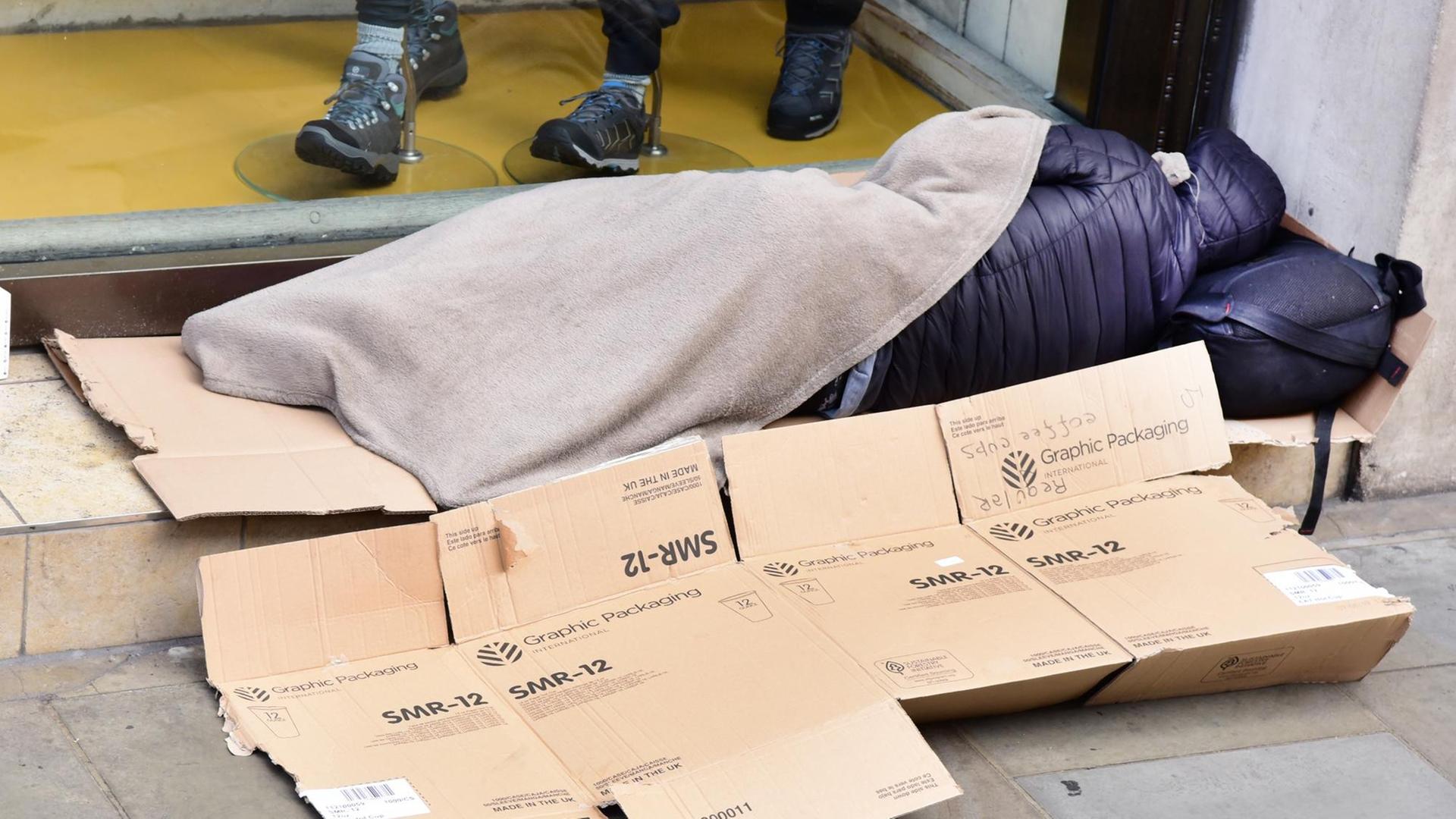 Vor einem Schaufenster liegt ein schlafender Obdachloser mit einer dünnen Decke auf Pappschachteln. Vor einem Schaufenster liegt ein schlafender Obdachloser mit einer dünnen Decke auf Pappschachteln.