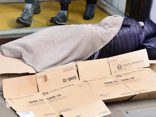 Vor einem Schaufenster liegt ein schlafender Obdachloser mit einer dünnen Decke auf Pappschachteln.