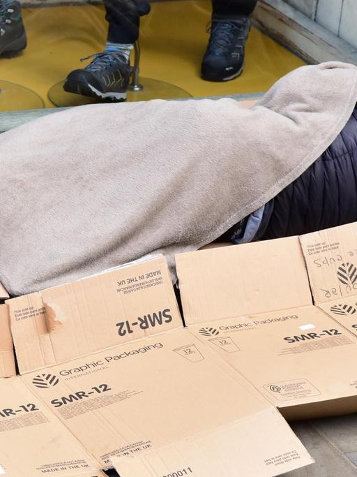 Vor einem Schaufenster liegt ein schlafender Obdachloser mit einer dünnen Decke auf Pappschachteln.