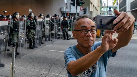 Ein älterer Mann macht ein Selfie von sich auf einer Straße in Hong Kong, mit Polizisten in voller Ausrüstung im Hintergrund, während der Demonstrationen gegen die Regierung.