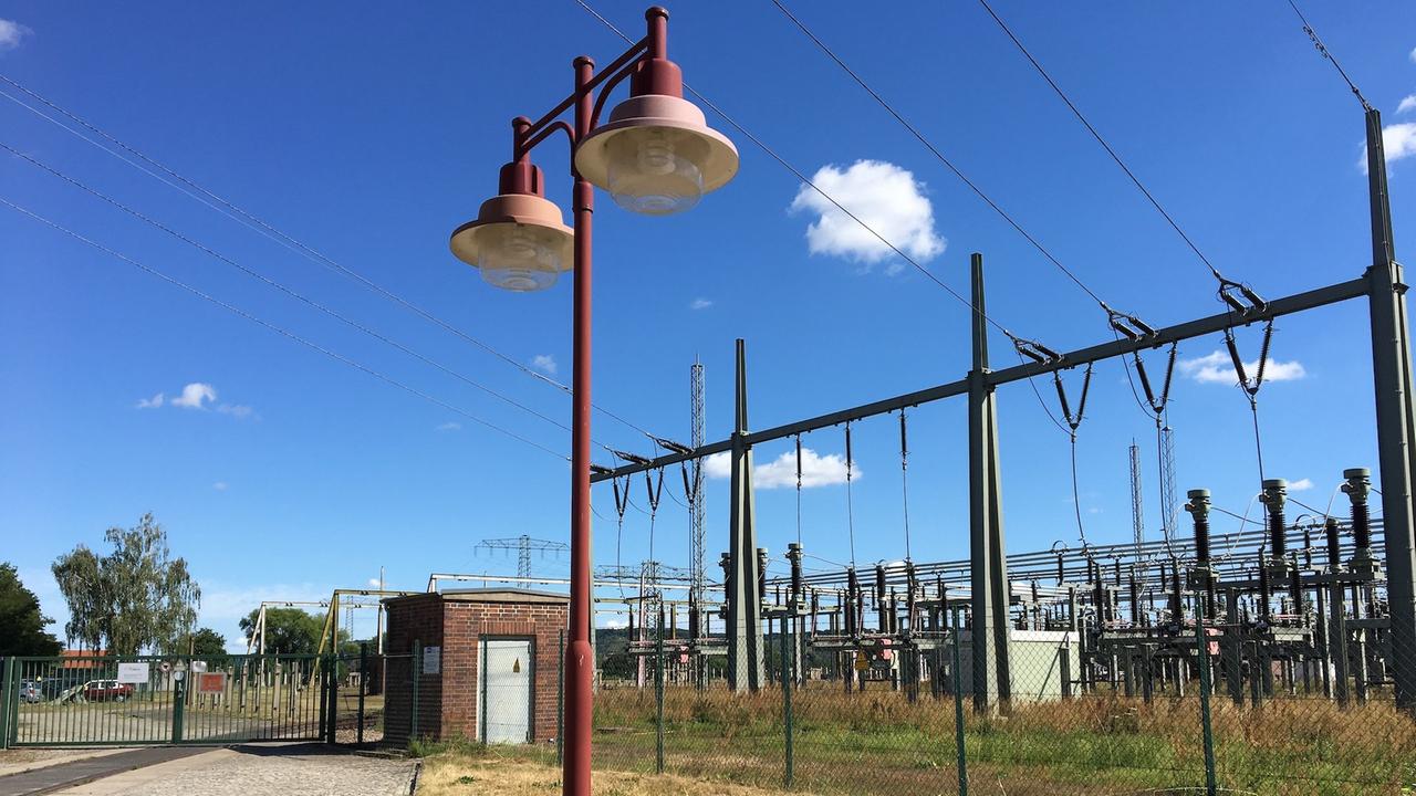 Ein Weg mit Kopfsteinpflaster führt zum Tor eines Industriegrundstücks. Im Vordergrund befindet sich eine verrostete Straßenlaterne, hinter dem Zaun ein Umspannwerk. Ein Weg mit Kopfsteinpflaster führt zum Tor eines Industriegrundstücks. Im Vordergrund befindet sich eine verrostete Straßenlaterne, hinter dem Zaun ein Umspannwerk.