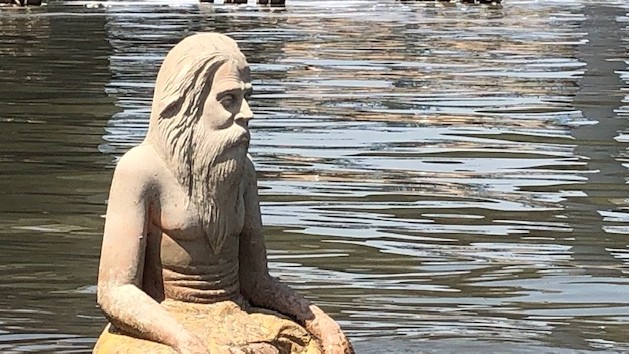 Eine helle männliche Steinfigur mit langen Haaren und Rauschebart sitzt im Schneidersitz in einem Gewässer.