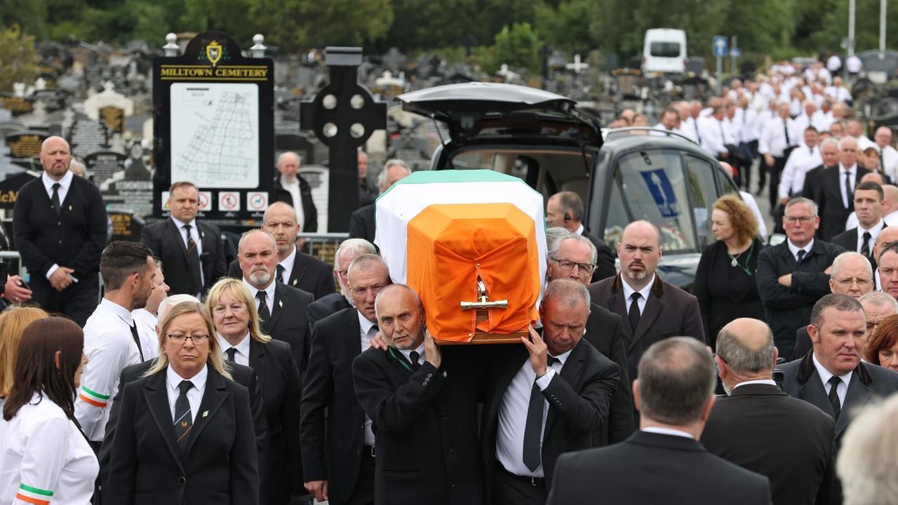 Ein Sarg, der in eine irische Flagge gehüllt ist, wird von Männern in dunklen Anzügen getragen. Dahinter zahlreiche Menschen, die der Prozession zum Friedhof folgen.