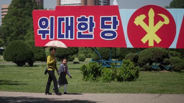 Eine Frau mit Sonnenschirm und ein Kind laufen durch einen Park, hinter ihnen ein Propagandaplakat.
