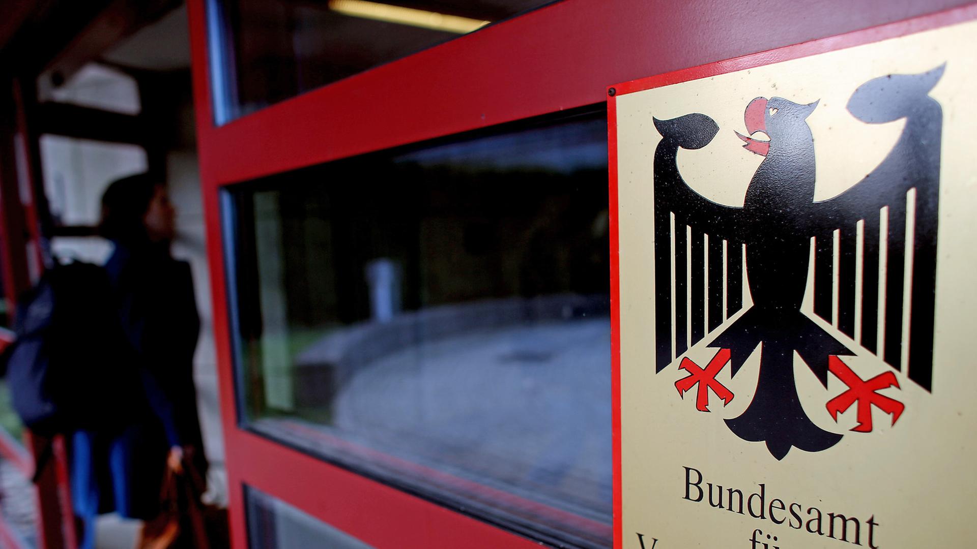 Schild des Bundesamts für Verfassungsschutz mit dem Bundesadler an einer Eingangstür des Gebäudes, durch die eine Frau geht. Schild des Bundesamts für Verfassungsschutz mit dem Bundesadler an einer Eingangstür des Gebäudes, durch die eine Frau geht.