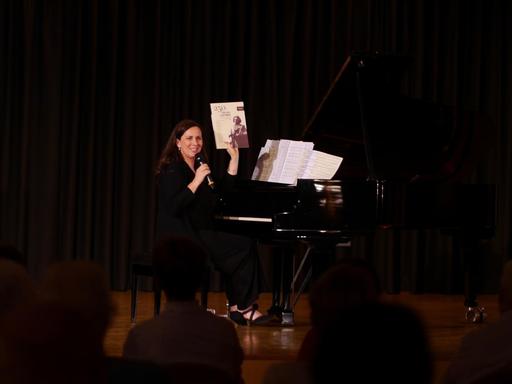 Pianistin Susanne Kessel sitzt mit einem Mikrofon in der Hand am Flügel. In der anderen Hand präsentiert sie eine Partiur ihres Projekts 250 piano pieces for Beethoven.