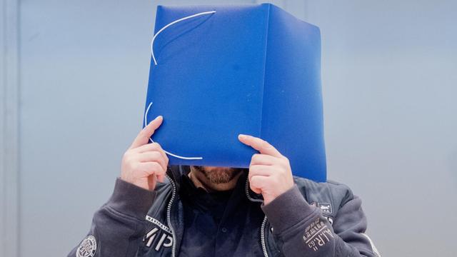 Der wegen vielfachen Mordes Angeklagte Niels Högel kommt in den Gerichtssaal und hält sich eine blaue Mappe vor das Gesicht