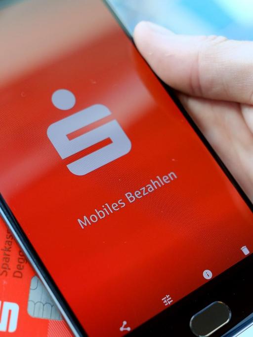 Ein Mann hält eine EC-Karte und ein Mobiltelefon, auf dem der Startbildschirm der Mobiles-Bezahlen-App der Sparkasse zu sehen ist, in der Hand. Knapp fünf Wochen nach Google sind die Sparkassen mit ihrem Smartphone-Bezahlsystem in Deutschland gestartet.