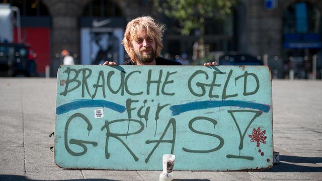 Ein Mann sitzt in Hamburg vor dem Hauptbahnhof und hält ein Schild mit der Aufschrift "Brauche Geld für Gras!"(Bild: Daniel Reinhardt/dpa )
