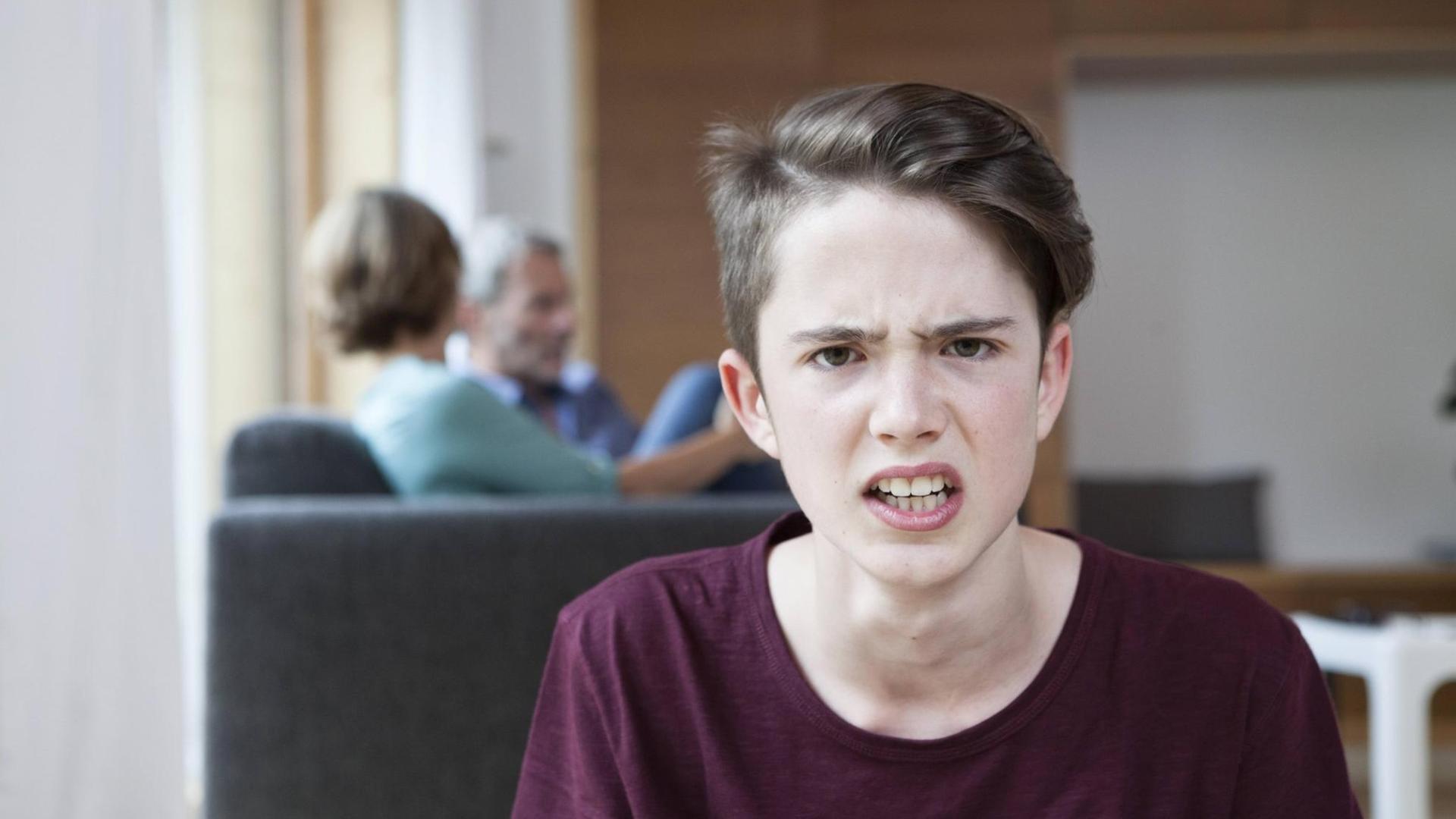 Ein wütender Junge, der in die Kamera guckt. Im Hintergrund sind die Eltern zu sehen. Ein wütender Junge, der in die Kamera guckt. Im Hintergrund sind die Eltern zu sehen.