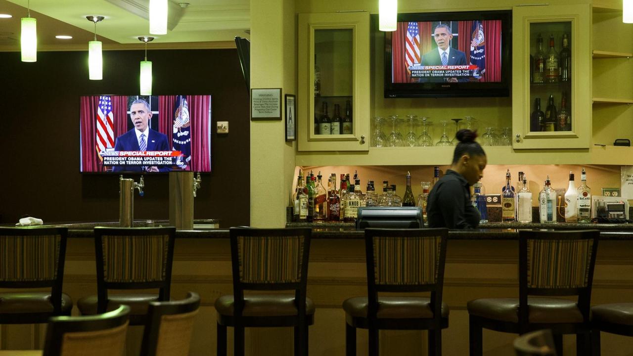Zwei Fernseher, auf denen Obamas Rede zu sehen ist, hängen hinter einer Bar mit weiblichem Barkeeper, der gerade beschäftigt ist.. Zwei Fernseher, auf denen Obamas Rede zu sehen ist, hängen hinter einer Bar mit weiblichem Barkeeper, der gerade beschäftigt ist..