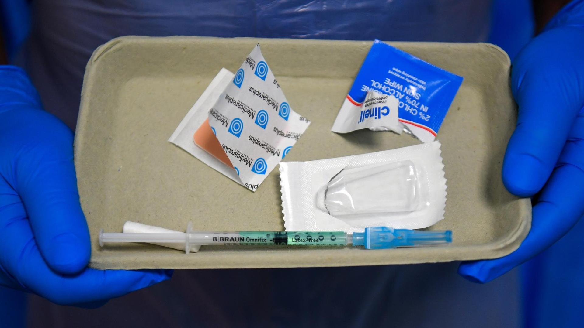 Eine Spritze und Pflaster liegen auf einem kleinen Tablett, das von zwei blau behandschuhten Händen getragen wird Eine Spritze und Pflaster liegen auf einem kleinen Tablett, das von zwei blau behandschuhten Händen getragen wird