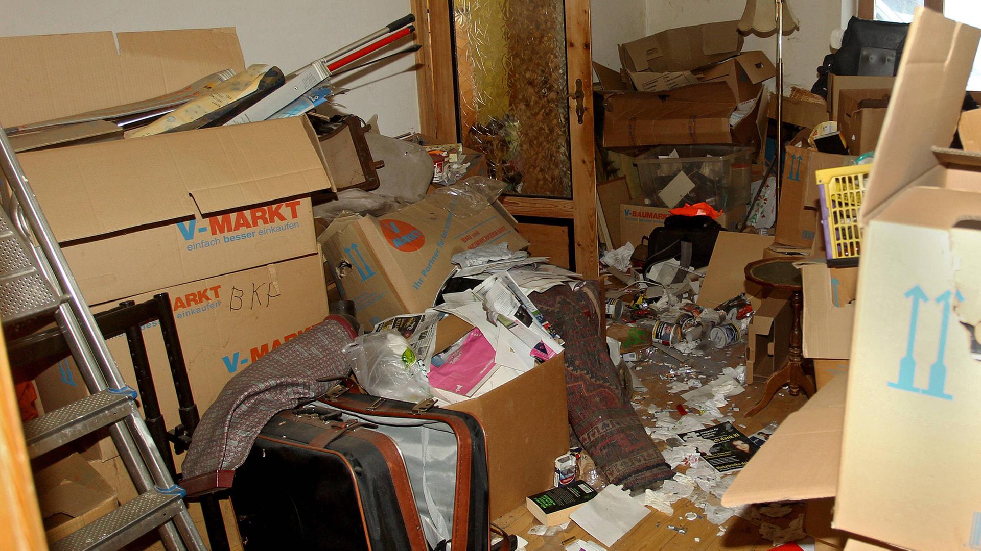 Eine vermüllte Wohnung: Kisten, eine Leiter, ein alter Koffer und viele weitere Gegenstände sind auf dem Boden zu sehen. Eine vermüllte Wohnung: Kisten, eine Leiter, ein alter Koffer und viele weitere Gegenstände sind auf dem Boden zu sehen.