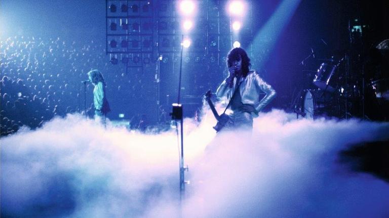 Die Rockband Led Zeppelin bei einem Live-Auftritt. Auf der Bühne ist viel Nebel, im Hintergrund ist eine große Menschenmenge zu sehen.