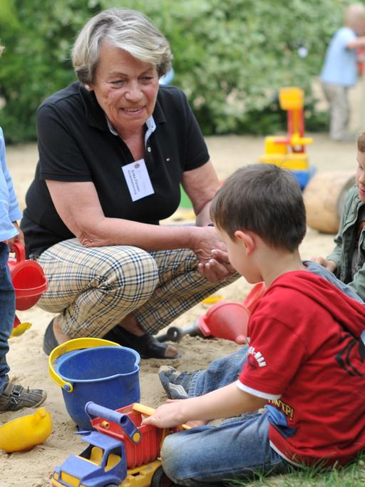 Eine Seniorin spielt im Garten des Mehrgenerationenhauses mit mehreren Kindern in einem Sandkasten.