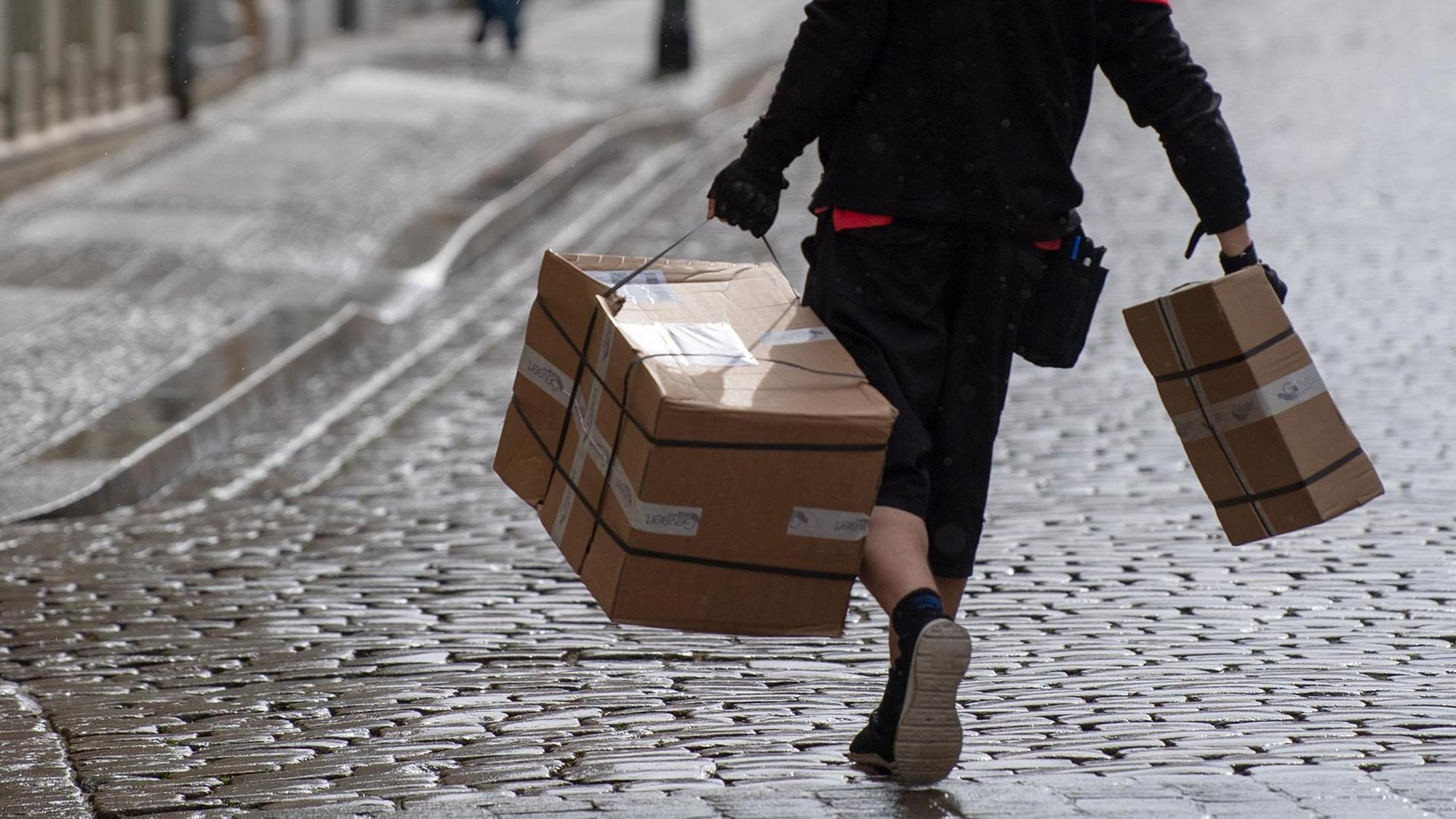 Ein Zusteller des Kurierdienstes dpd trägt an beiden Seiten jeweils ein schweres Paket durch die Straße. Ein Zusteller des Kurierdienstes dpd trägt an beiden Seiten jeweils ein schweres Paket durch die Straße.