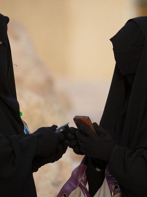 Zwei Frauen, voll verschleiert mit der traditonellen Abaya, tippen in Riad in Saudi-Arabien auf ihren Handys.