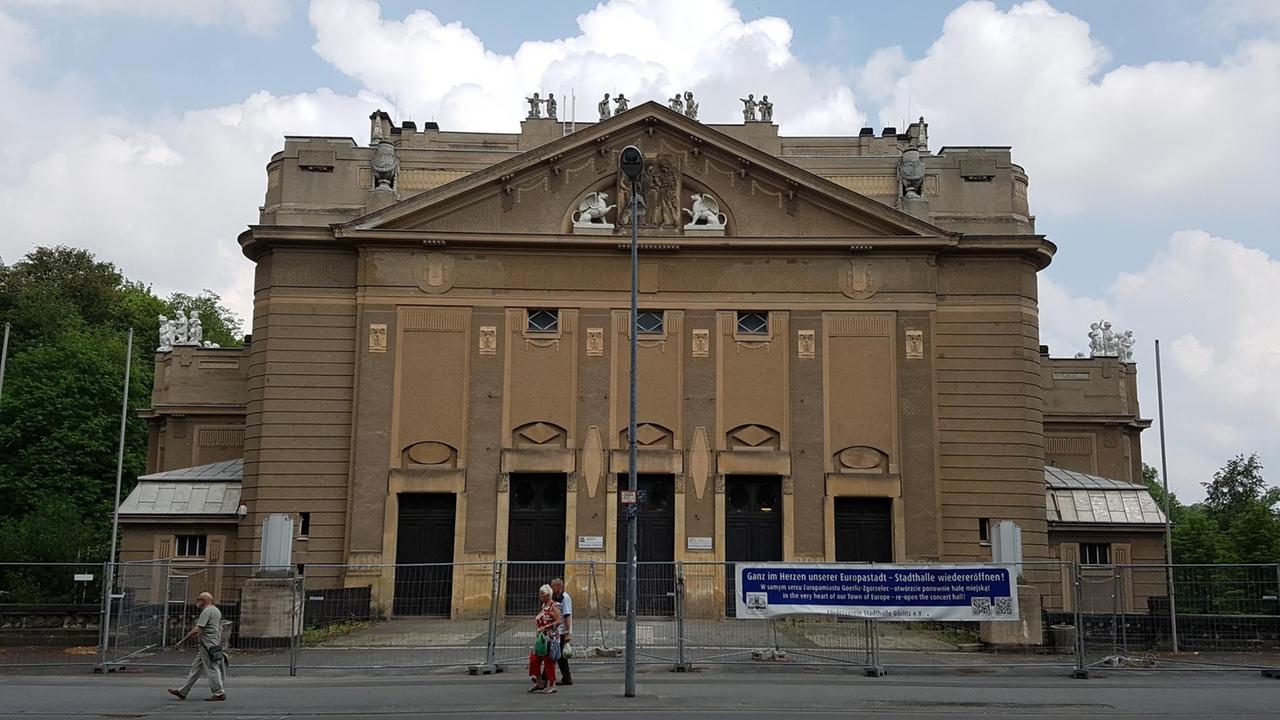 Vor einem großen braunen Gebäude mit Bauzaun, gehen Menschen von rechts nach links vorbei. Vor einem großen braunen Gebäude mit Bauzaun, gehen Menschen von rechts nach links vorbei.