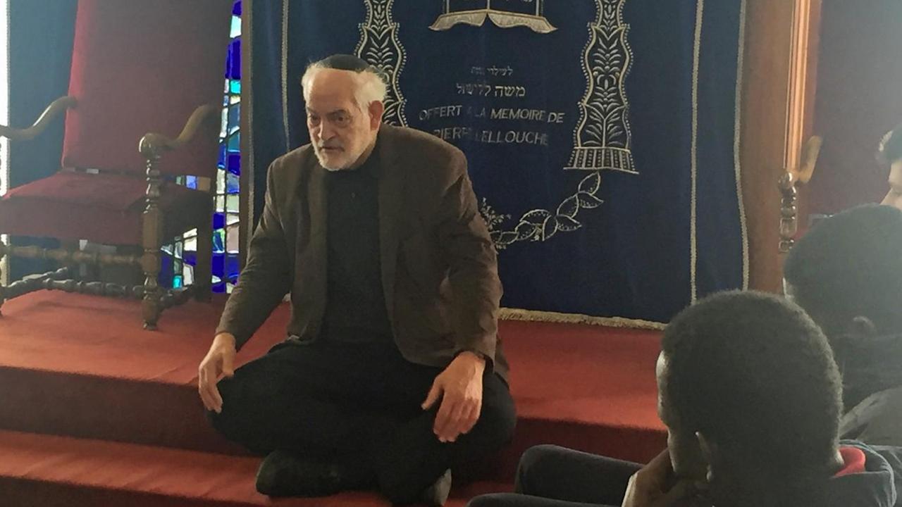 Der Rabbiner Michel Serfaty sitzt in der Synagoge auf einer Stufe und spricht mit muslimischen Jugendlichen