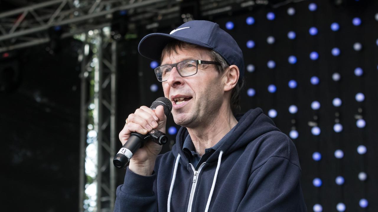 Ein Mann im blauen Kaputzenpullover und blauer Kappe steht auf einer Bühne. Er spricht in ein Mikrofon. Ein Mann im blauen Kaputzenpullover und blauer Kappe steht auf einer Bühne. Er spricht in ein Mikrofon.
