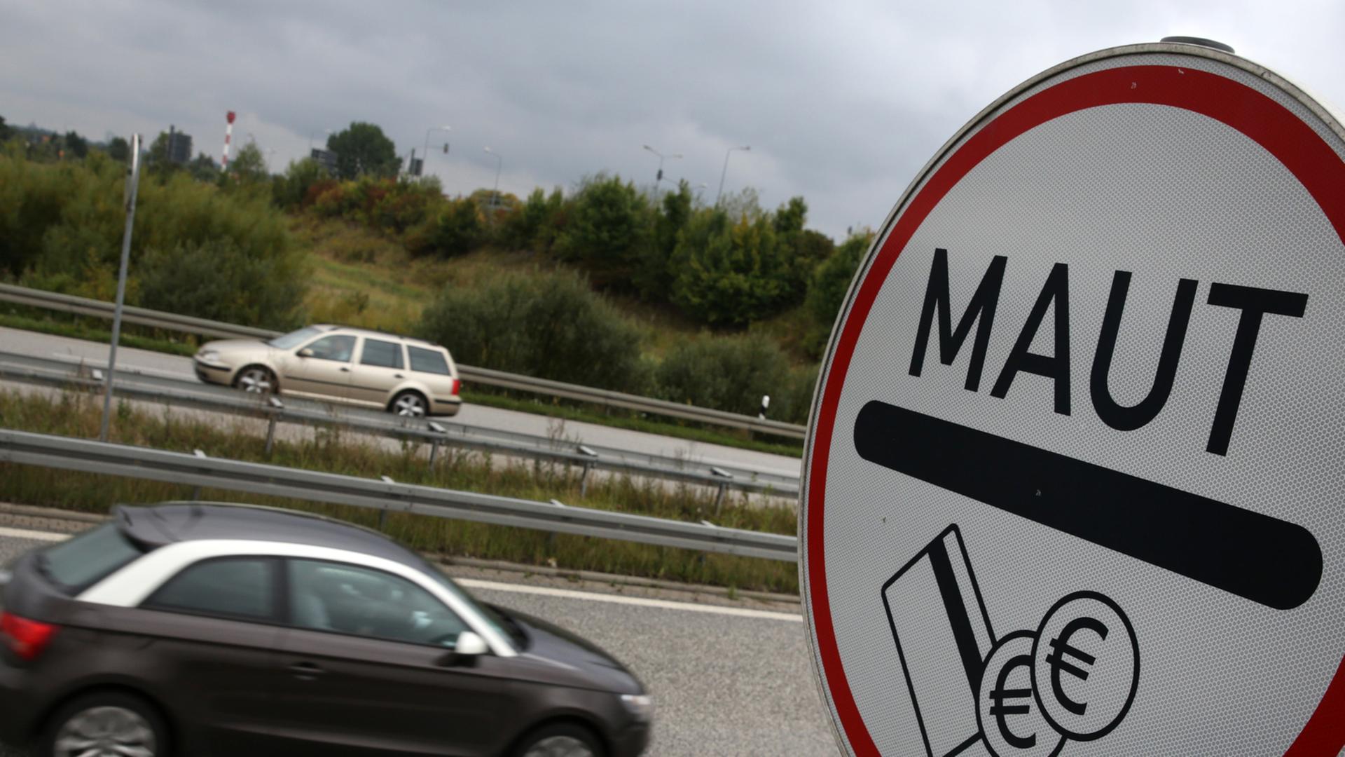 Ein Schild mit der Aufschrift "Maut" steht an einer Autobahn, im Hintergrund fahren Autos vorbei. Ein Schild mit der Aufschrift "Maut" steht an einer Autobahn, im Hintergrund fahren Autos vorbei.