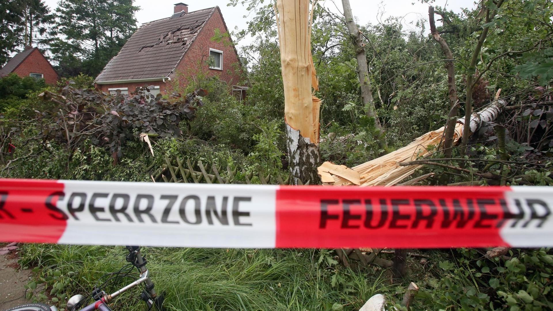 Hinter einem Absperrband der Feuerwehr sind abgeknickte Bäume und ein beschädigtes Dach zu sehen. Hinter einem Absperrband der Feuerwehr sind abgeknickte Bäume und ein beschädigtes Dach zu sehen.