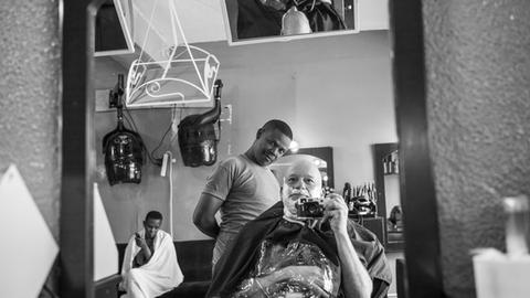 Der Photojournalist Karsten Thielker fotografiert sich in einem Spiegel selbst, während ihm ein Barbier in Kigali den Bart stutzt. Das Bild ist schwarz/weiß.