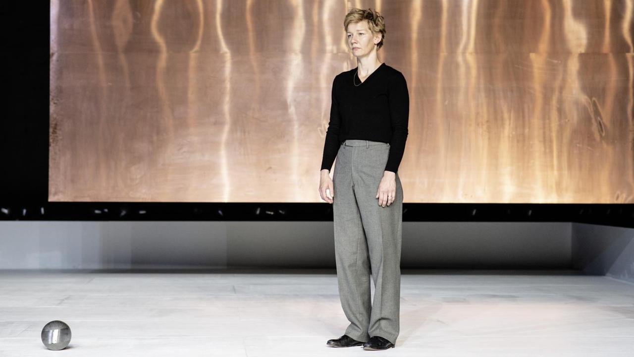 Eine junge Frau steht traurig vor einem abstrakten Hintergrund auf einer Theaterbühne. Vor ihr liegt eine metallisch schimmernde Kugel. Eine junge Frau steht traurig vor einem abstrakten Hintergrund auf einer Theaterbühne. Vor ihr liegt eine metallisch schimmernde Kugel.