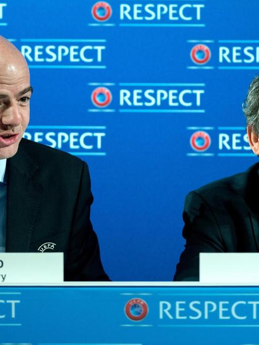 Infantino und Platini sitzen bei einer pressekonferenz und lächeln.