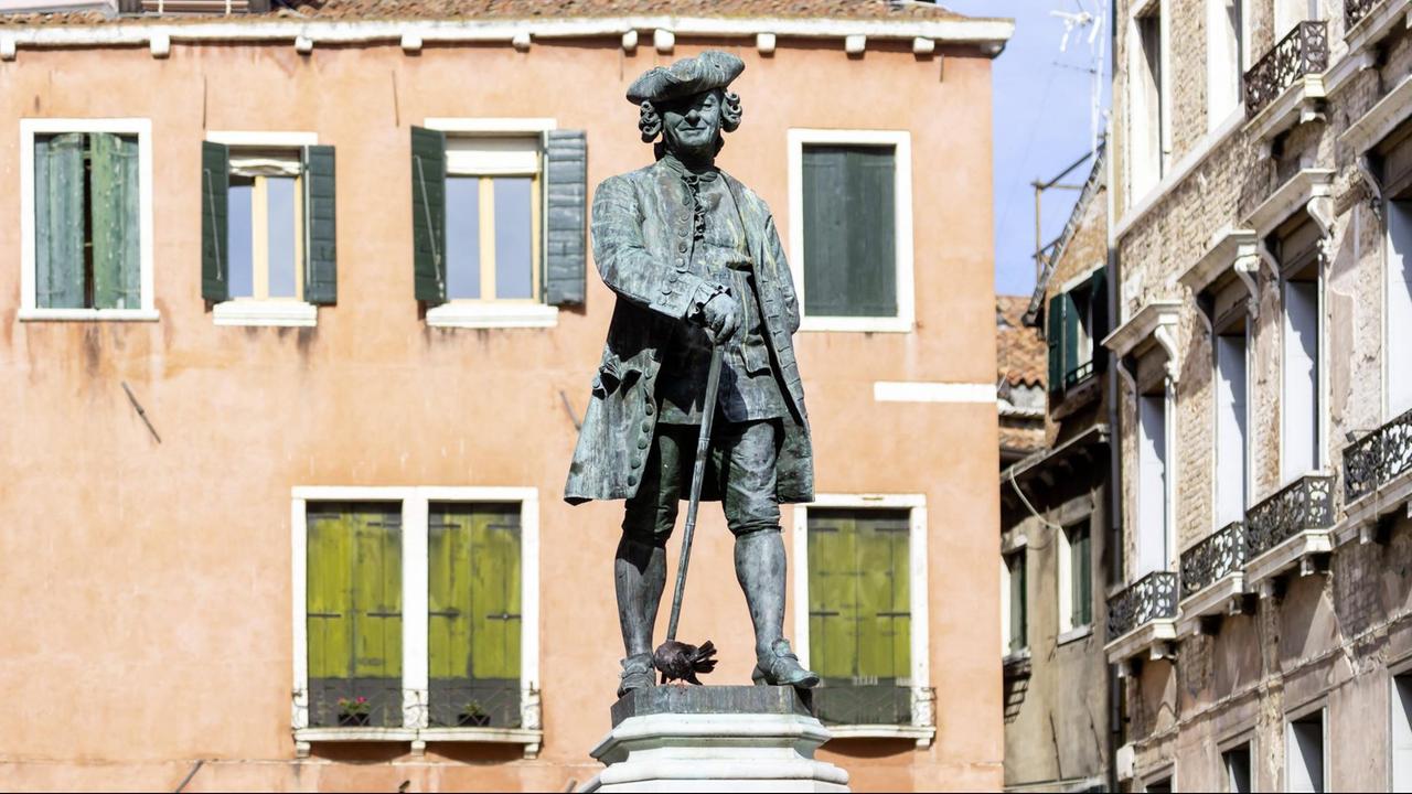 Lächelnde Statue mit Hut und Spazierstock vor den typisch venezianischen Häusern.