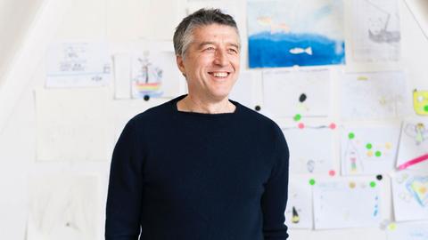 Der Unternehmer Andreas Murkudis in einem schwarzen Pullover vor einer Wand, an die Zeichnungen gepinnt sind.