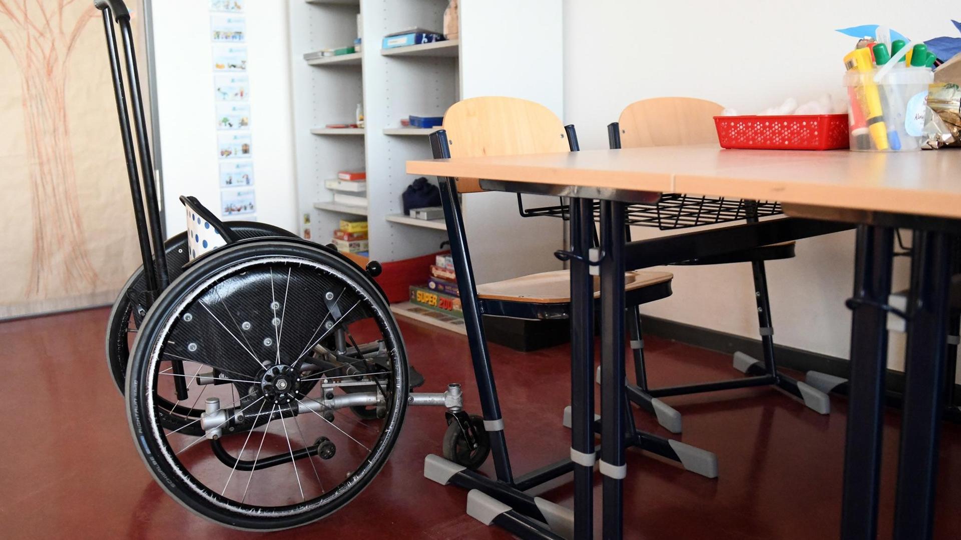 An einem Gruppentisch im Klassenraum stehen zwei Holzstühle und ein Kinderrollstuhl. An einem Gruppentisch im Klassenraum stehen zwei Holzstühle und ein Kinderrollstuhl.