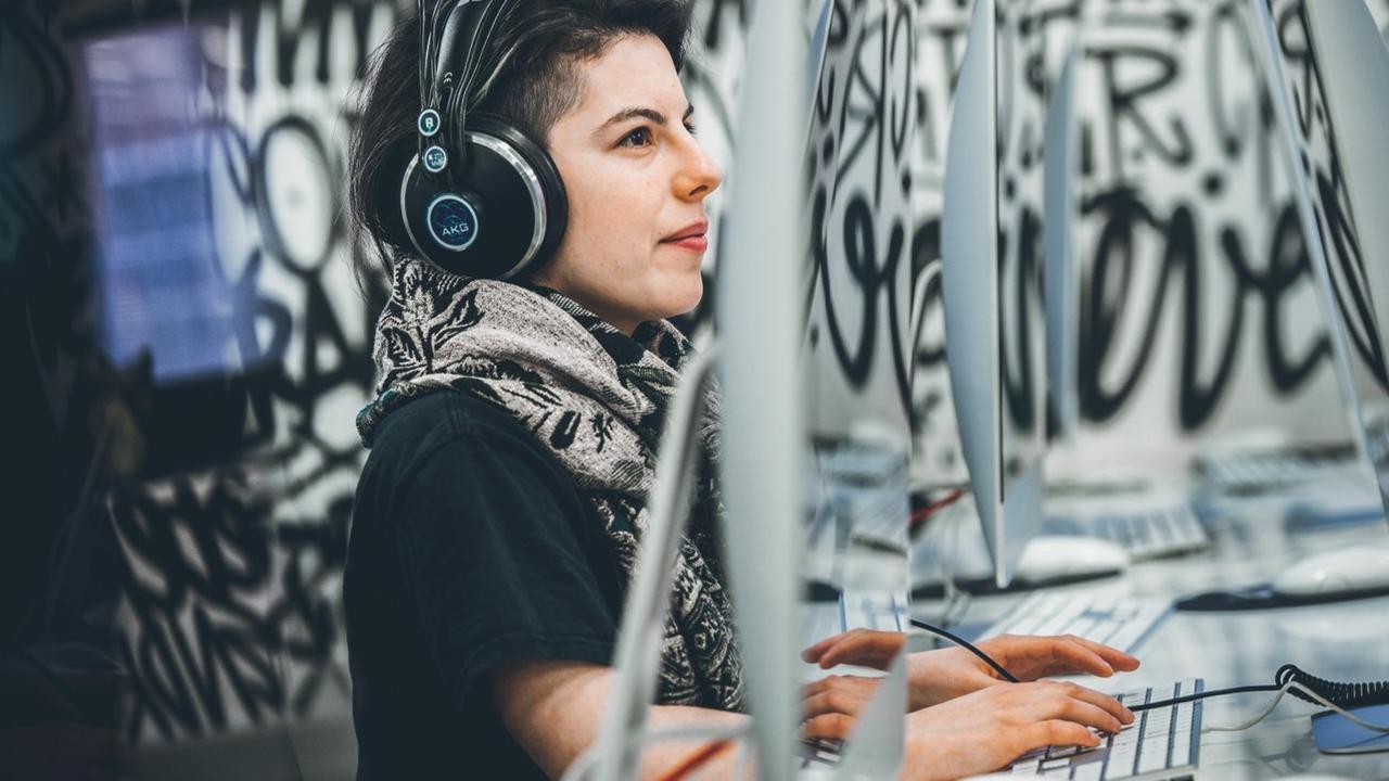 Eine junge Frau mit kurzen Haaren und roten Lippen trägt große schwarze Kopfhörer bei der Arbeit am Computer. Im Hintergrund sind Graffiti zu sehen. Eine junge Frau mit kurzen Haaren und roten Lippen trägt große schwarze Kopfhörer bei der Arbeit am Computer. Im Hintergrund sind Graffiti zu sehen.