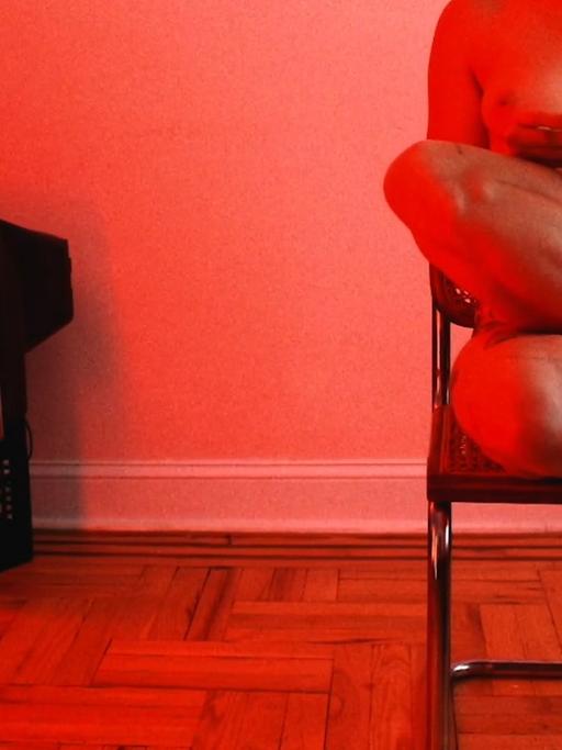 Szene aus dem Kurzfilm "Marriage Story": In einem in rotes Licht getauchten Raum steht links ein alter Fernseher, rechts sitze eine nackte Frau auf einem Stuhl und liest.