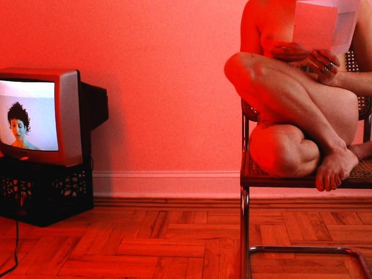 Szene aus dem Kurzfilm "Marriage Story": In einem in rotes Licht getauchten Raum steht links ein alter Fernseher, rechts sitze eine nackte Frau auf einem Stuhl und liest.