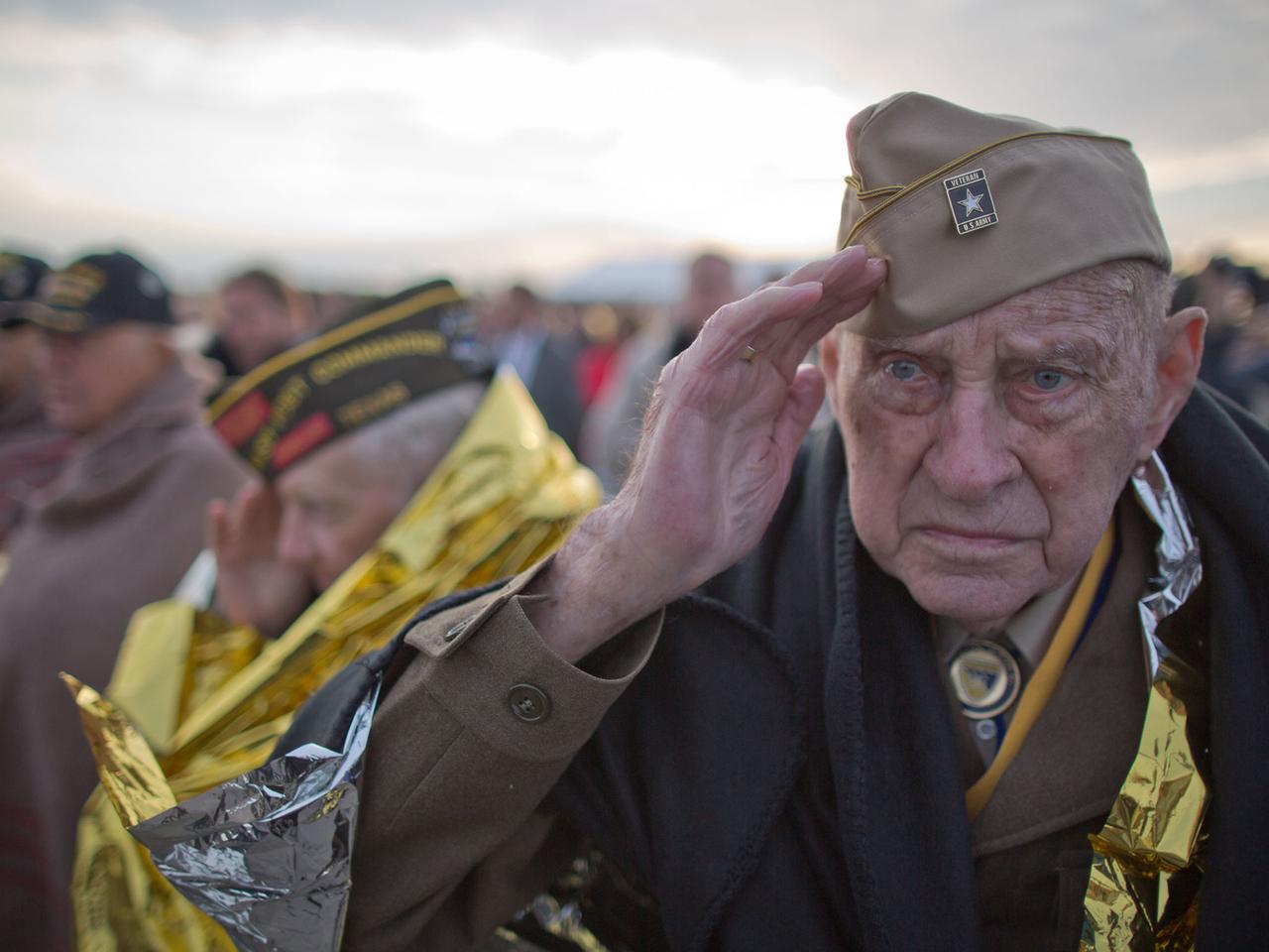 Weltkriegsveteran Raymond Sylvester aus den USA salutiert am 05.06.2014 während einer Gedenkveranstaltung am Utah Beach in der Normandie in Frankreich, eingehüllt in eine Wärmedecke.