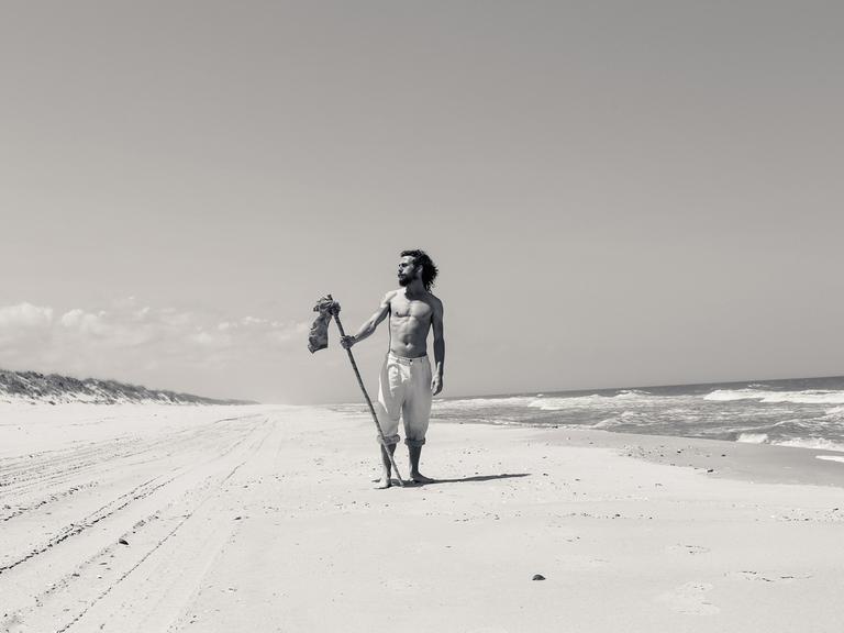 Ein Mann mit langen Haaren und Bart steht, nur mit einer kurzen Hose bekleidet, an einem Meeresstand. Er hält einen Stock in der Hand und schaut in die Ferne.